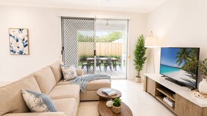 Ein Wohnzimmer mit beiger Couch, Couchtisch, Fernseher auf Holzfuß und einer Schiebetür, die auf eine Terrasse im Freien führt.