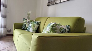 A green sofa with leaf-patterned pillows, a white vase with a single white flower, and a wall with a textured artwork.