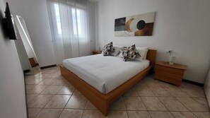 A bedroom with a bed, a nightstand, a wall-mounted TV, and a window with curtains.