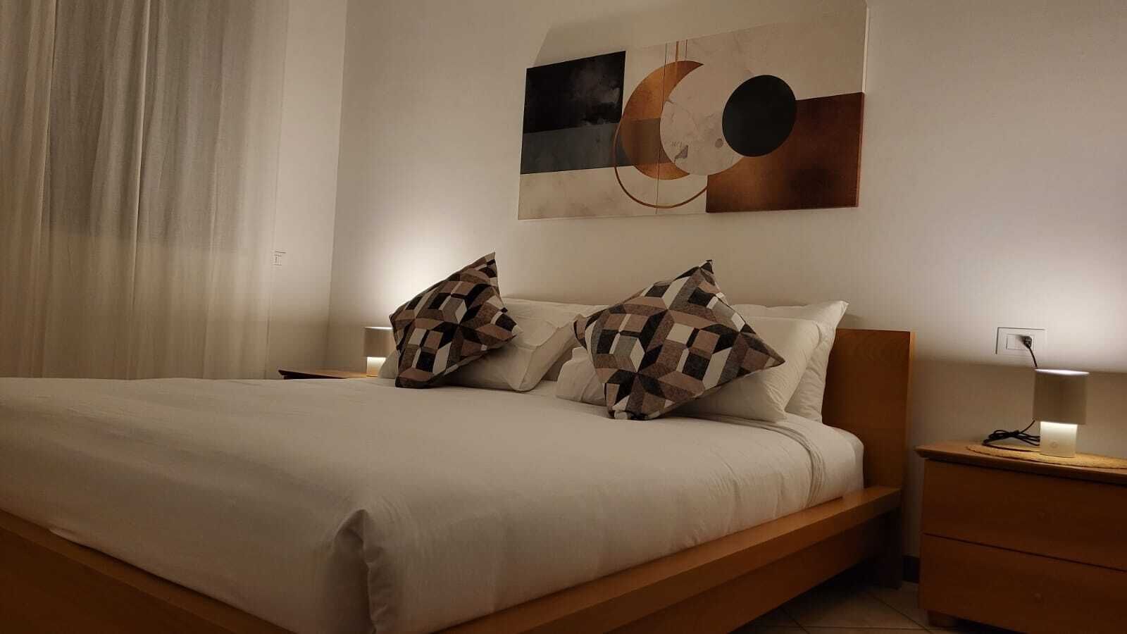 A neatly made bed with two patterned pillows, a bedside table with a lamp, and a wall with abstract artwork.