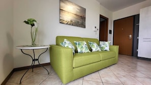 Ein grünes Sofa mit gemusterten Kissen, ein kleiner runder Tisch mit einer Pflanze und ein Gemälde an der Wand.