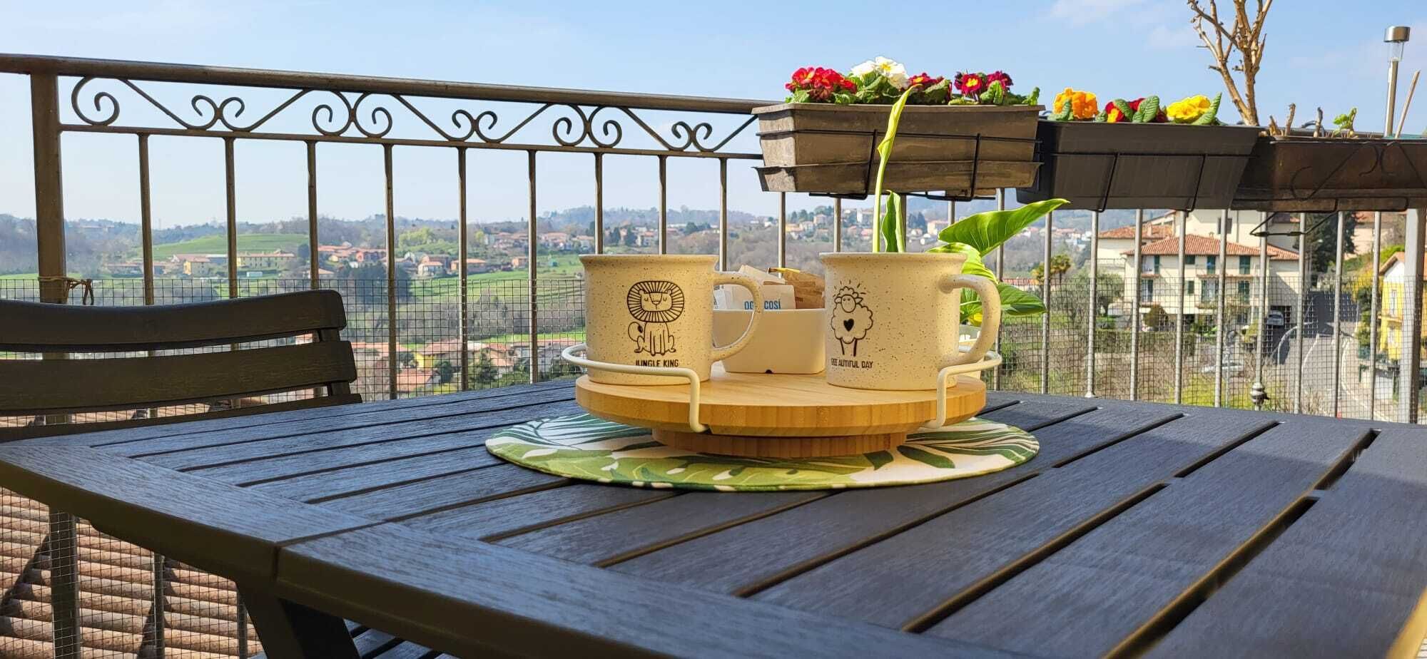 Ein Holztisch mit einer Ablage, zwei Tassen und einem Blumentopf auf einem Balkon mit Metallgeländer und Blick auf eine Stadt.