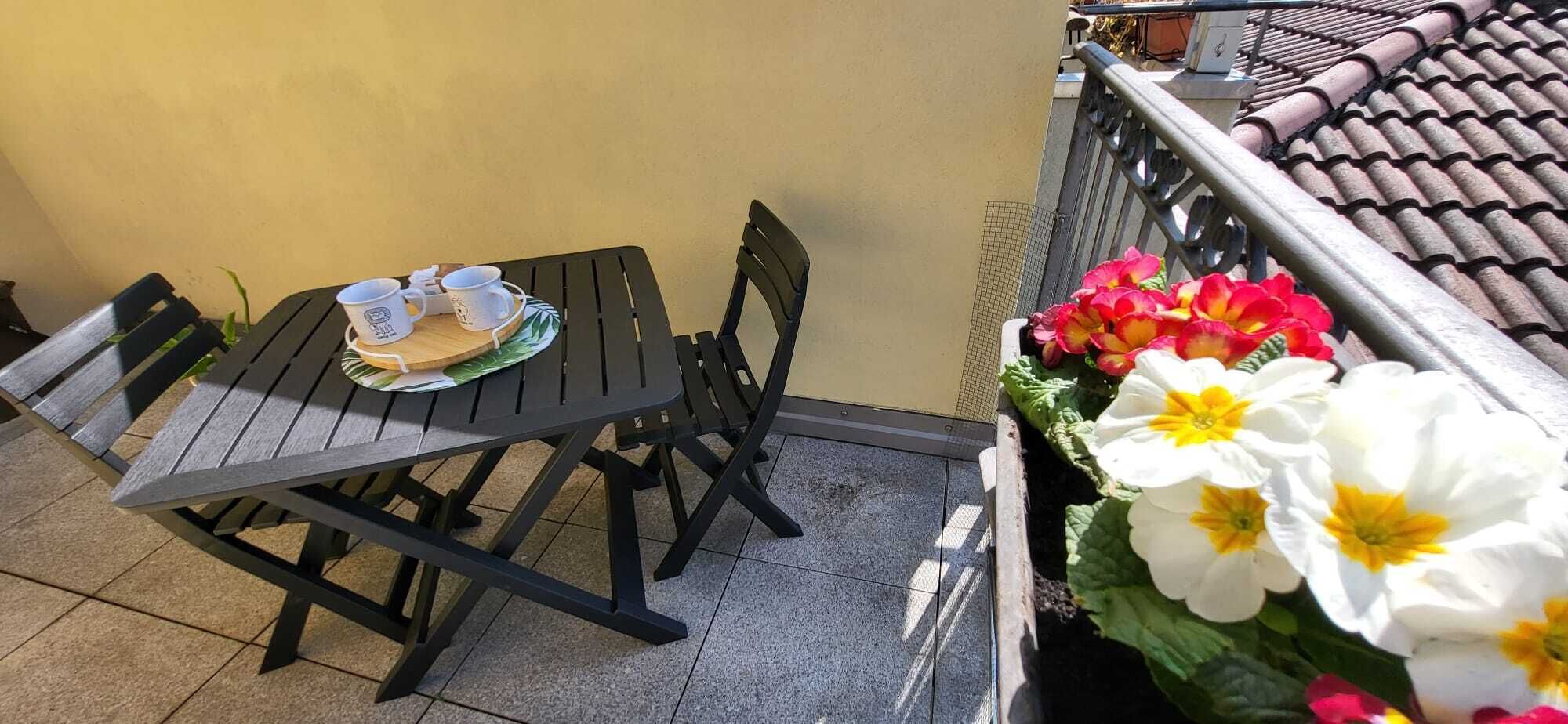 Un balcone con tavolo e sedie, una piccola pianta fiorita e un pavimento piastrellato.