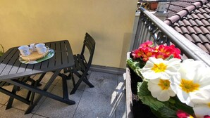 A balcony with a table and chairs, a small plant with flowers, and a tiled floor.