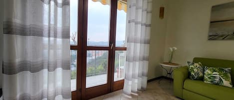 Una habitación con un sofá verde, una mesita redonda con un florero y vistas a través de un ventanal.