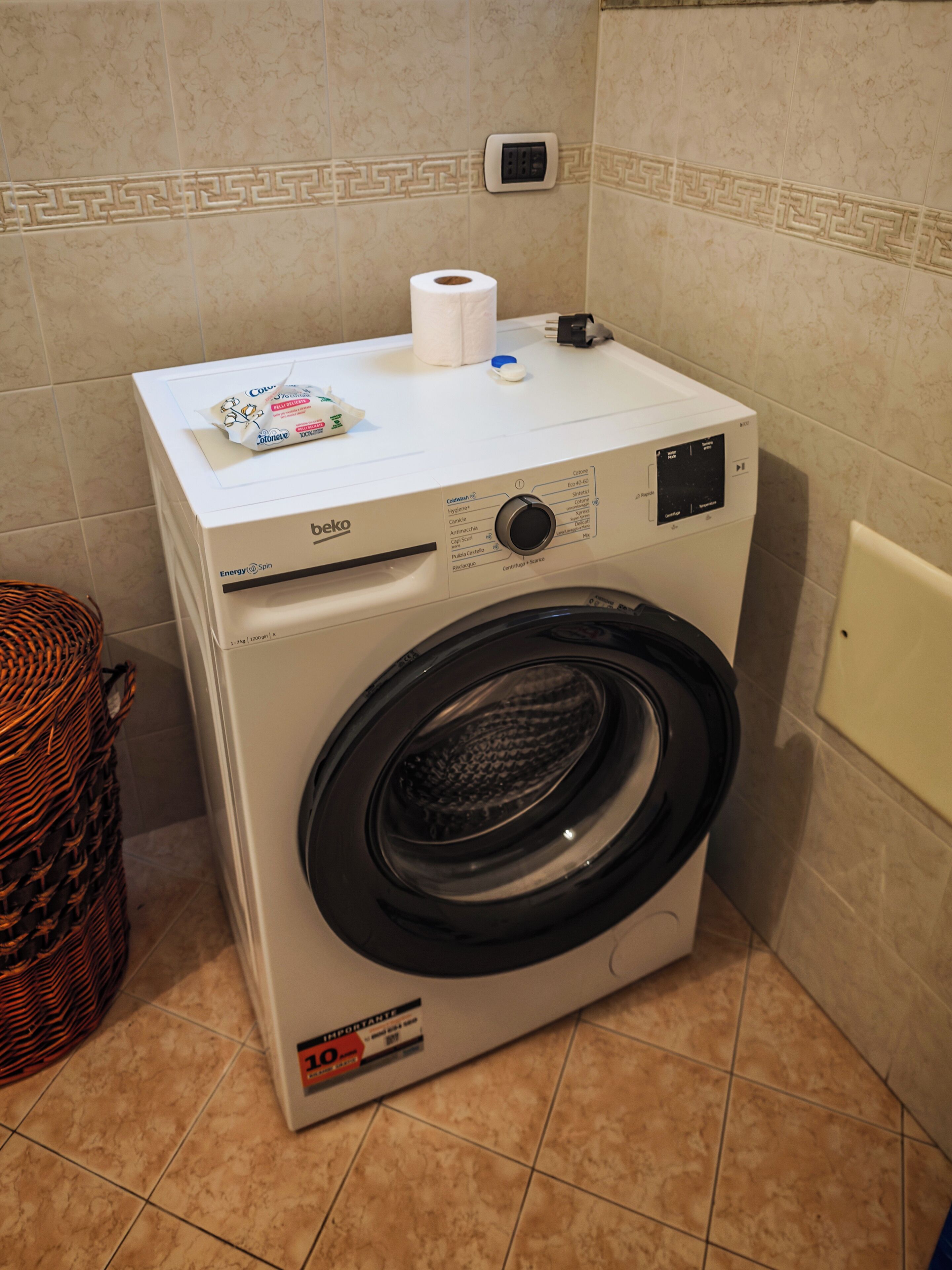 A front-loading washing machine with a control panel, a power outlet, and a wall-mounted electrical socket.
