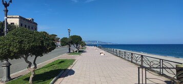 Un sendero pavimentado a lo largo de la costa, con baranda de metal, árboles y un edificio al fondo.