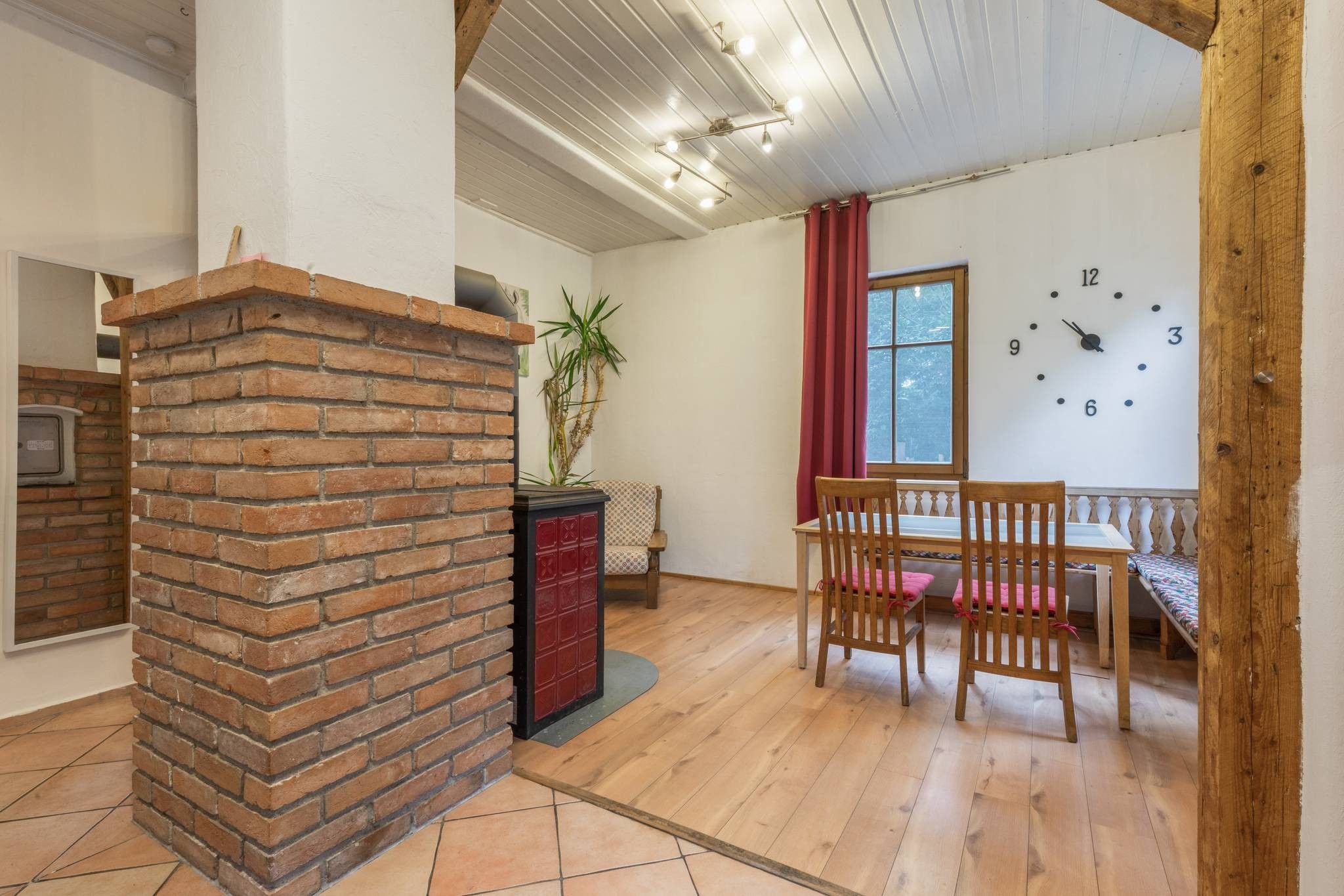 Una habitación con chimenea de ladrillo, una mesa de comedor de madera con sillas, un reloj en la pared y una ventana con cortinas.