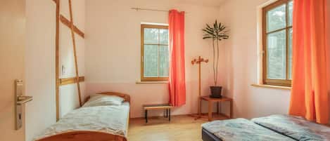 Een slaapkamer met twee bedden, een houten vloer, een raam met gordijnen, een kamerplant en een hangende lamp.