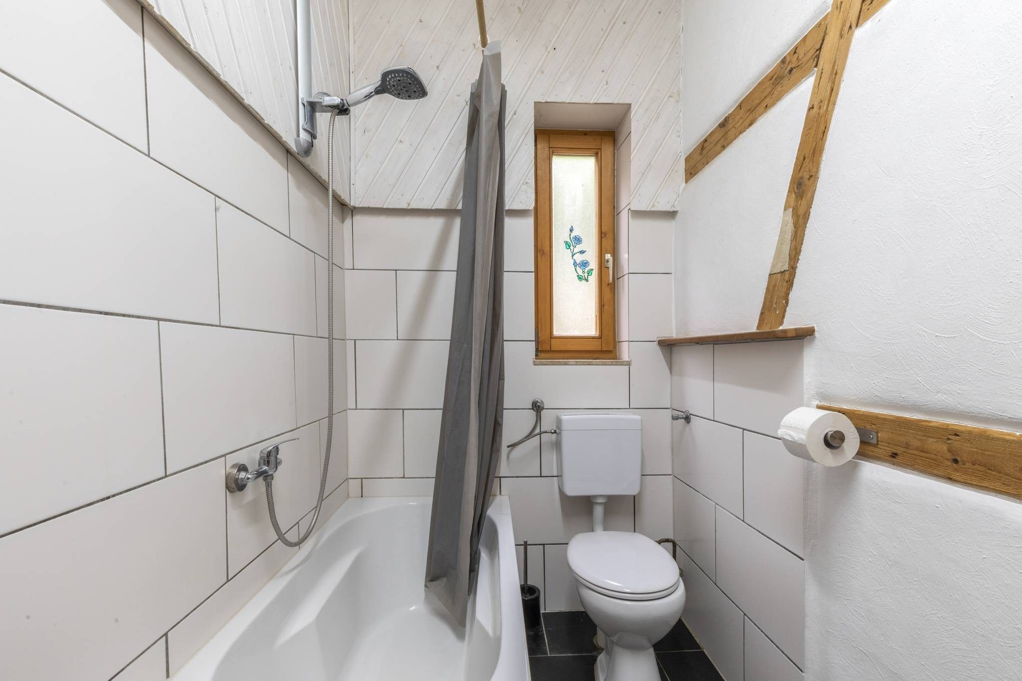 Ein kleines, kompaktes Badezimmer mit weißen Fliesen, einer weißen Toilette, einer Dusche inklusive Handbrause und einem Fenster mit Holzrahmen.
