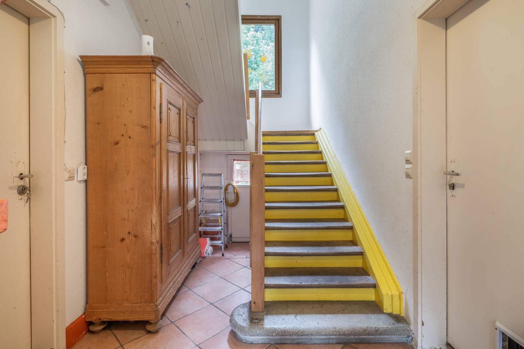 A staircase with yellow steps leading to a landing, a wooden cabinet, and a window with a view of trees.