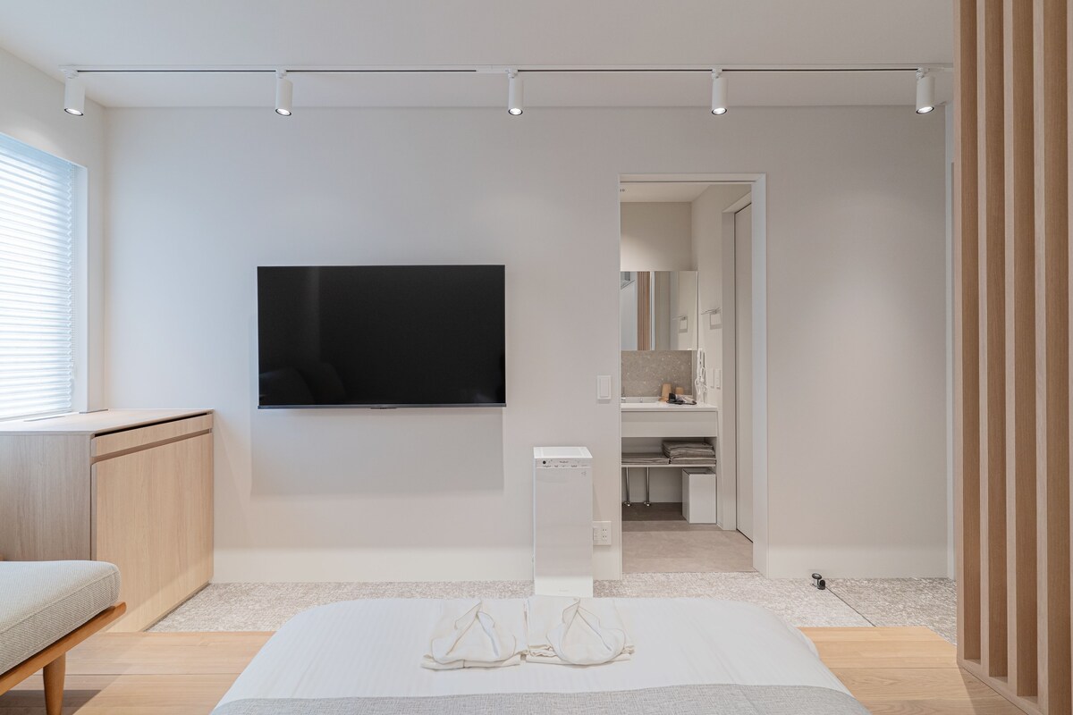 A modern, minimalist room with a flat-screen TV mounted on the wall, a wooden cabinet, a white sofa, and a small white table.
