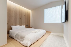 Un dormitorio con cabecera de madera, cama con ropa de cama blanca, mesita de noche y persianas en la ventana.
