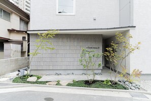 A building with a corrugated metal facade, a small tree in the foreground, and a paved area with rocks and greenery. - Giraffe Nakasu haku (Fukuoka)