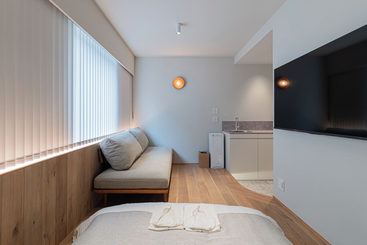 A compact hotel room with a bed, a seating area, a kitchenette, and a television mounted on the wall.