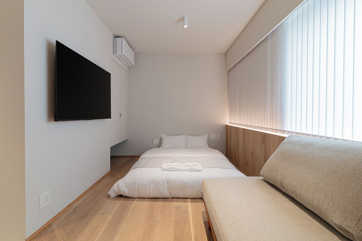 A modern bedroom with a bed, a flat-screen TV mounted on the wall, a bedside table, and a window with blinds.