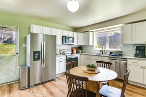 Een keuken met witte kastjes, roestvrijstalen apparatuur, een houten eettafel en een raam met uitzicht op een achtertuin.