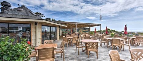 An outdoor patio with wooden tables and chairs, a covered seating area, and a view of the sea.