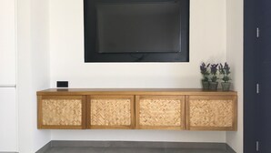 A flat-screen TV mounted on a wall above a wooden console with a woven back panel. - Elsa Modern Apartments (Pogonia)