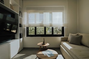 A modern living room with a flat-screen TV, a sofa, a small round table with a lamp, and a window with blinds. - Elsa Modern Apartments (Pogonia)