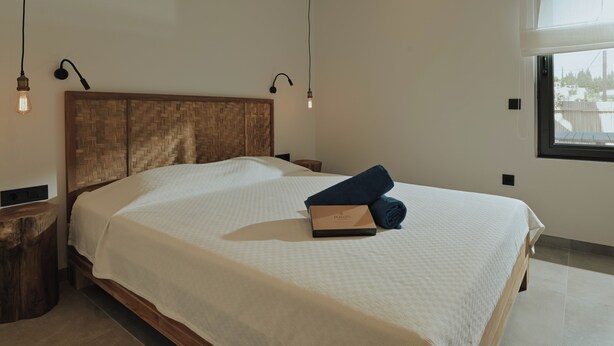 A neatly made bed with a wooden headboard, a bedside table, a lamp, and a window with a view. - Elsa Modern Apartments (Pogonia)