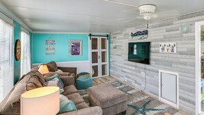 Ein Wohnzimmer mit einer Couch, einem Hocker, einer Lampe und einem an der Wand montierten Fernseher.