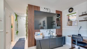 Ein modernes Wohnzimmer mit einem großen Flachbildfernseher, einem dunklen Holztisch mit Stuhl, einem blauen Teppich und Dekorationen auf Regalen.