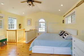 Um quarto com uma cama, mesa de cabeceira, cômoda, janela com persianas, ventilador de teto e escada que leva a um andar superior.