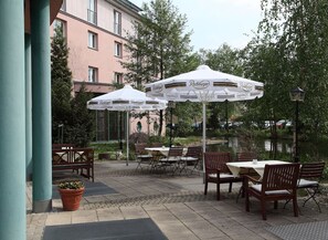 Ein Außenbereich mit Terrasse, Tischen, Stühlen unter weißen Sonnenschirmen, einer Holzbank und einem Blumentopf.