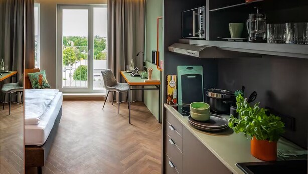 Habitación de hotel con cama, escritorio, silla, kitchenette y ventanas amplias.