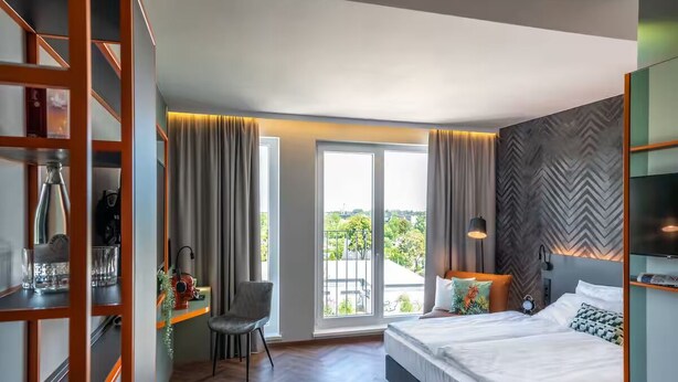 Een moderne hotelkamer met een groot bed, een bureau met stoel, een balkon met gordijnen en een wand met een herringbone patroon.