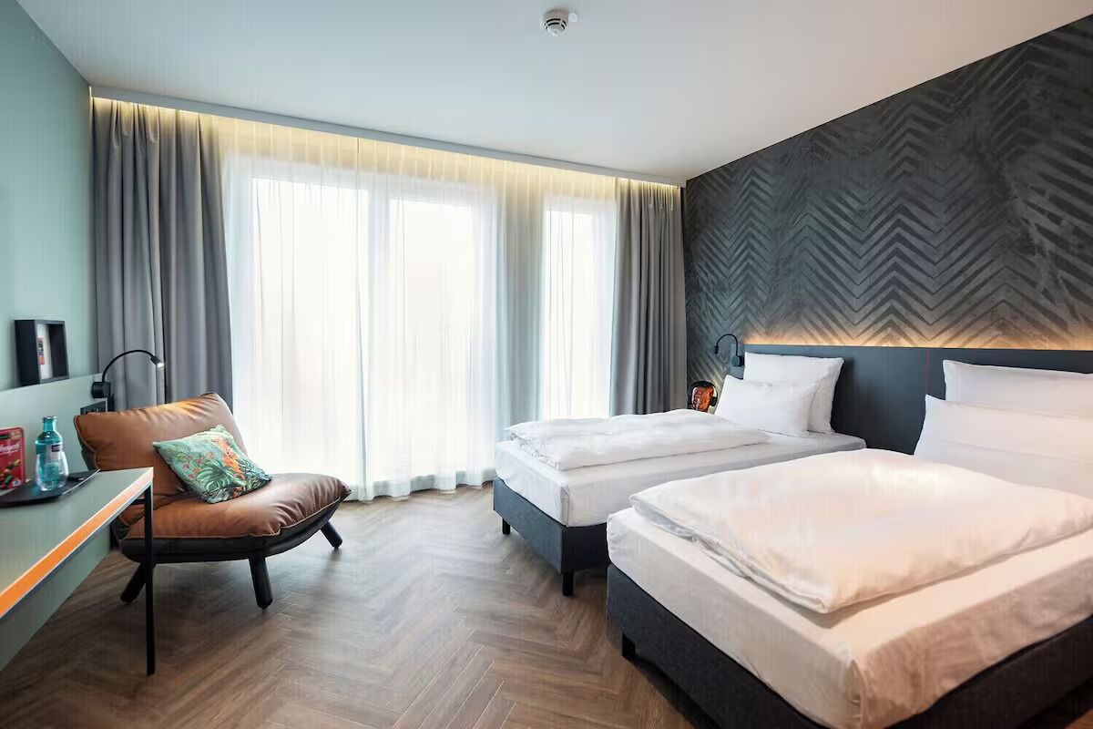 Quarto de hotel com duas camas, uma escrivaninha, uma cadeira e uma janela grande com cortinas.