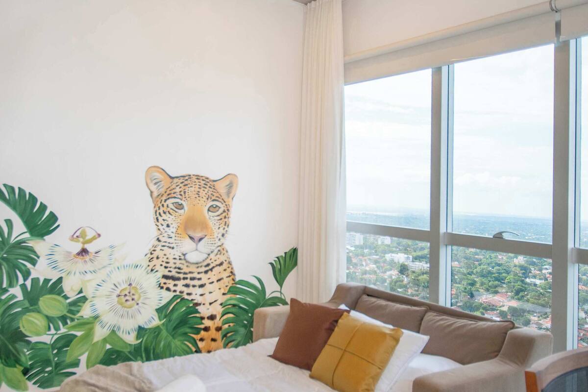 Une chambre avec une grande fenêtre, un lit, un canapé et un tableau représentant un léopard.