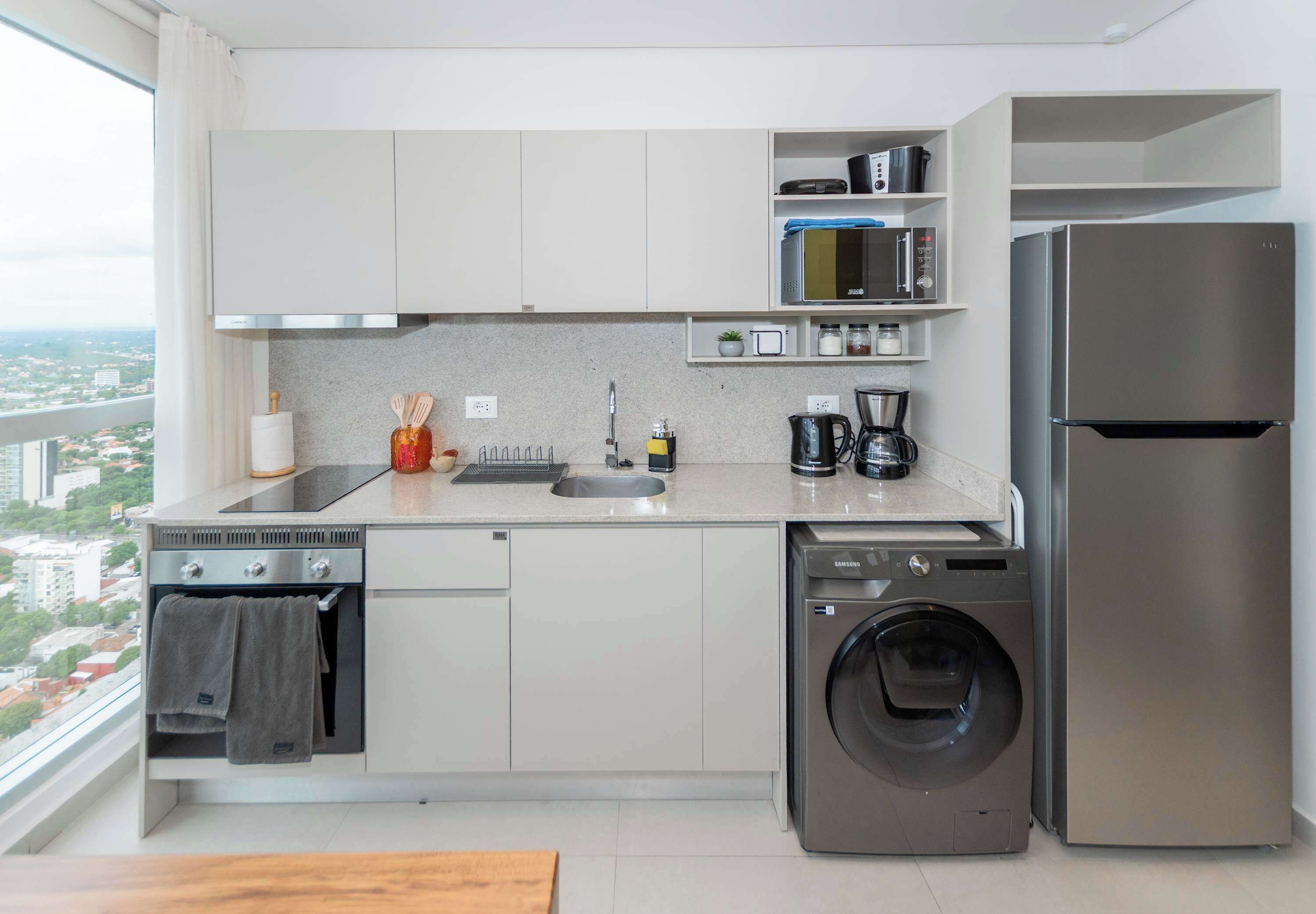 A modern kitchen with a built-in washer and dryer, a stainless steel refrigerator, and a window offering a city view.