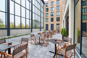 Ein Außenbereich mit Terrasse, Tischen, Stühlen, großen Fenstern und einem Gebäude im Hintergrund.