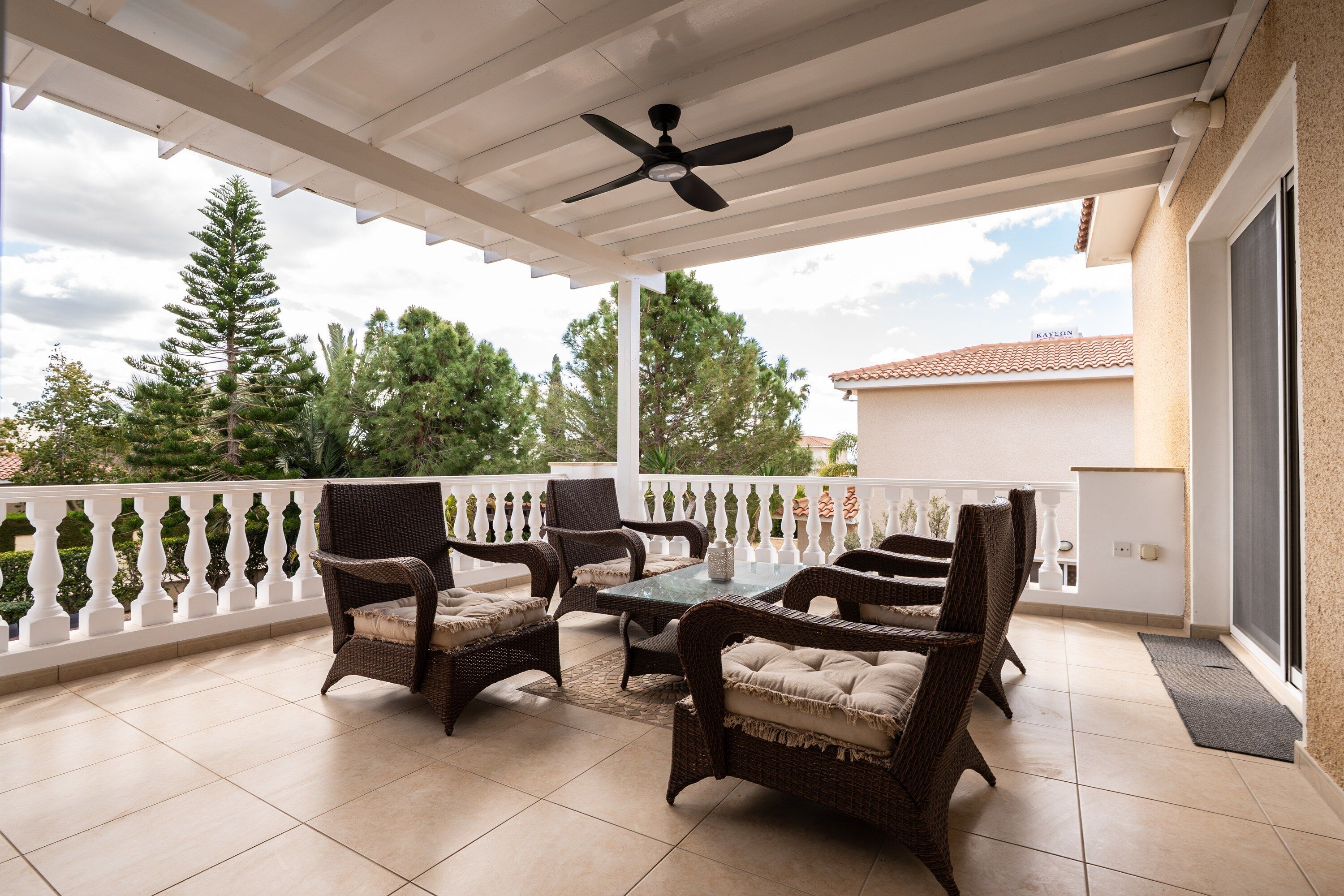 Une terrasse couverte, aménagée avec des meubles en osier, dotée d’un ventilateur de plafond et offrant une vue sur les arbres et les bâtiments.
