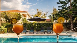 Blick über den Pool mit zwei Getränken, einem Pool und einem Gebäude mit Säulen im Hintergrund.