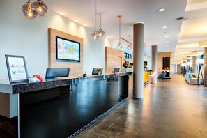 Ein modernes Hotel-Rezeptionslayout mit einem großen Empfangstresen, verschiedenen Sitzgelegenheiten und einem großen Bildschirm, der eine Präsentation zeigt.