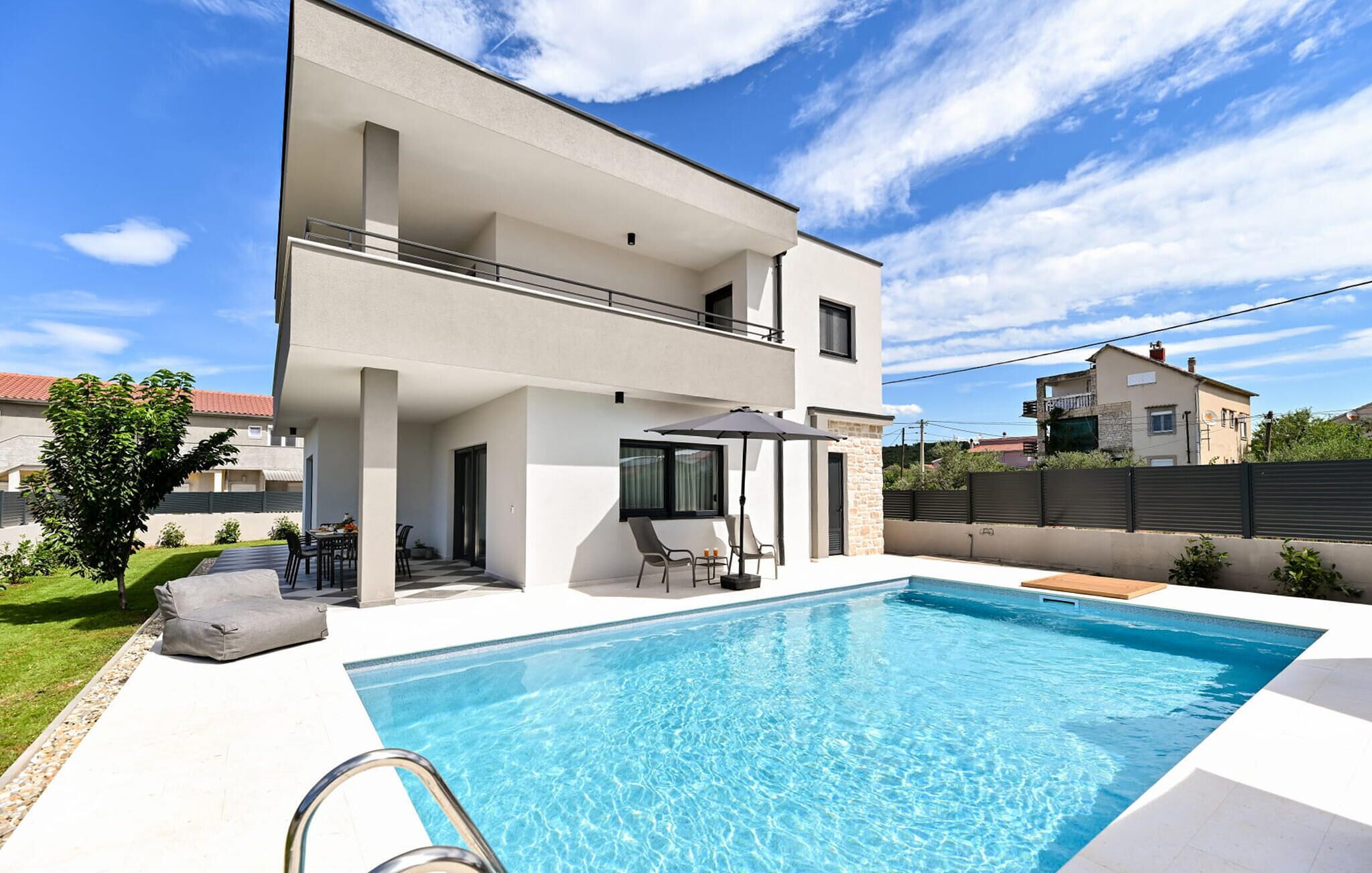Una casa moderna de dos pisos con piscina, mobiliario exterior y vistas a casas vecinas.