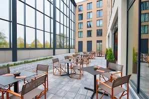 Ein Außenbereich mit Terrasse, Tischen, Stühlen, großen Fenstern und einem Gebäude im Hintergrund.
