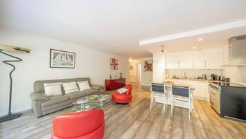 A modern living room with a sofa, red chair, glass coffee table, and a kitchen area with white cabinets and appliances.