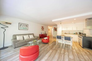 Una sala moderna con sofá, sillón rojo, mesa de centro de vidrio y una cocina con armarios y electrodomésticos blancos.