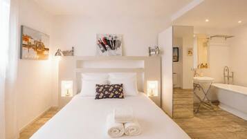 Une chambre d’hôtel avec un grand lit, une table de chevet, une lampe fixée au mur, un tableau et une salle de bain visible à travers une porte ouverte.