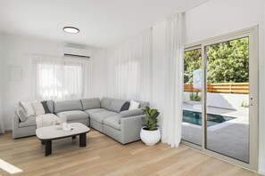 A modern living room with a grey sectional sofa, a white coffee table, a potted plant, and a sliding glass door leading to a pool area.