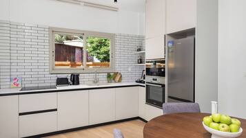 A modern kitchen with white cabinets, a stainless steel refrigerator, a wooden dining table, and a bowl of green apples.