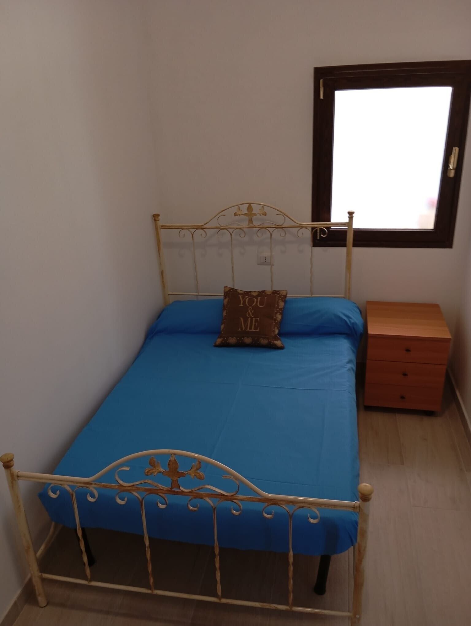 Un lit simple avec un matelas bleu, une tête de lit en métal, une table de chevet avec des tiroirs et une fenêtre au-dessus du lit.