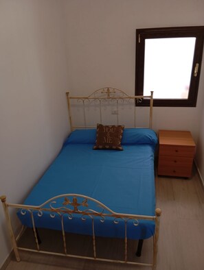 Un lit simple avec un matelas bleu, une tête de lit en métal, une table de chevet avec des tiroirs et une fenêtre au-dessus du lit.