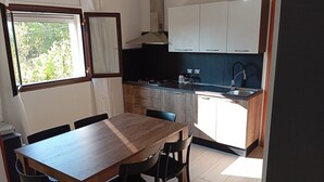 Een kleine keuken met een houten tafel en stoelen, een raam met gordijnen, en witte keukenkastjes.