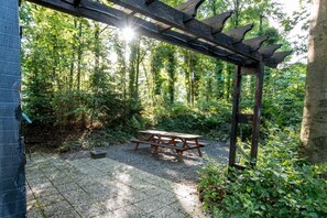 A covered picnic area with a table and benches, surrounded by a forested area.