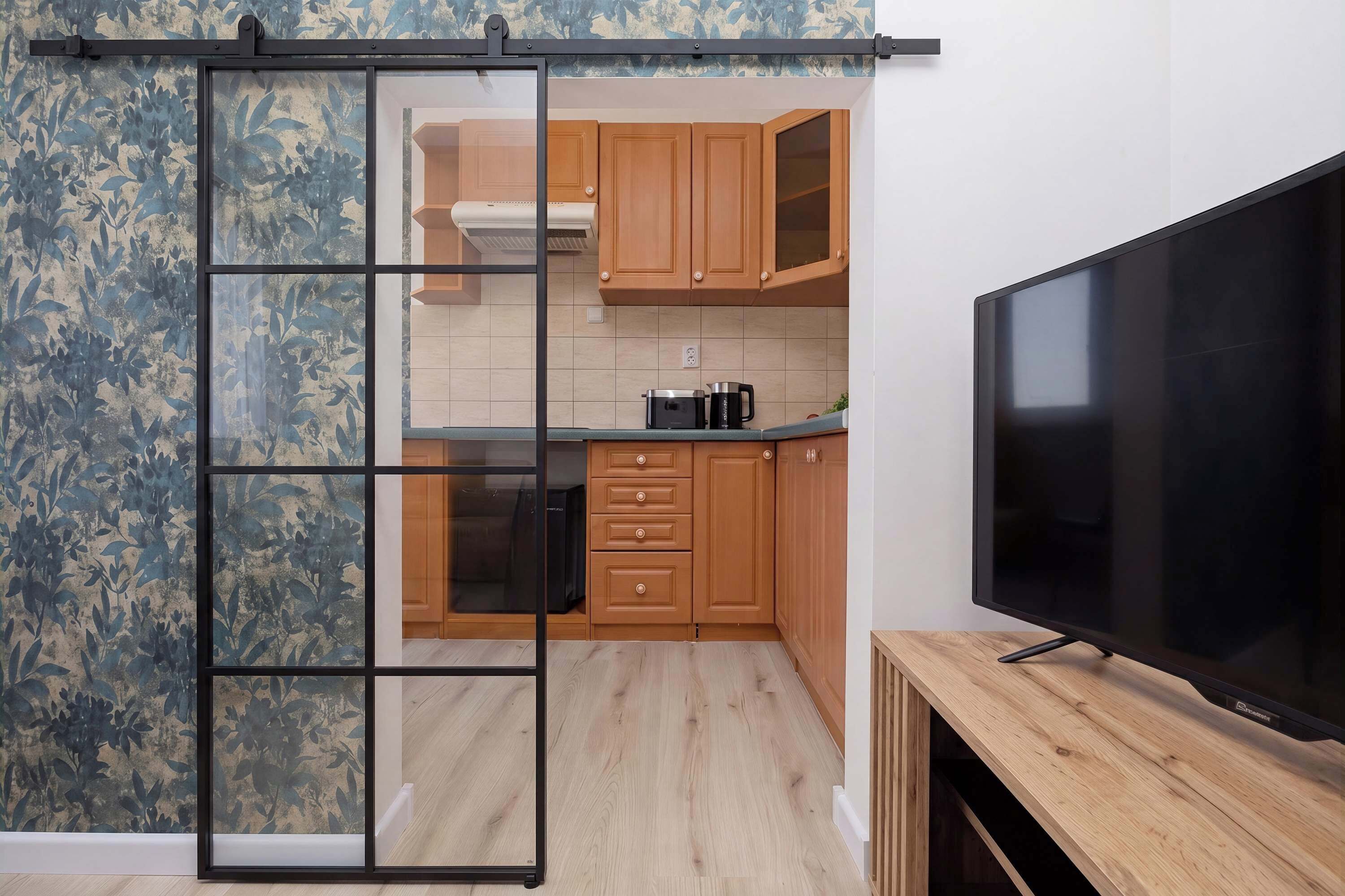 Une cuisine moderne avec des meubles en bois, un téléviseur noir posé sur un meuble en bois, et une porte-fenêtre coulissante donnant sur une autre pièce.